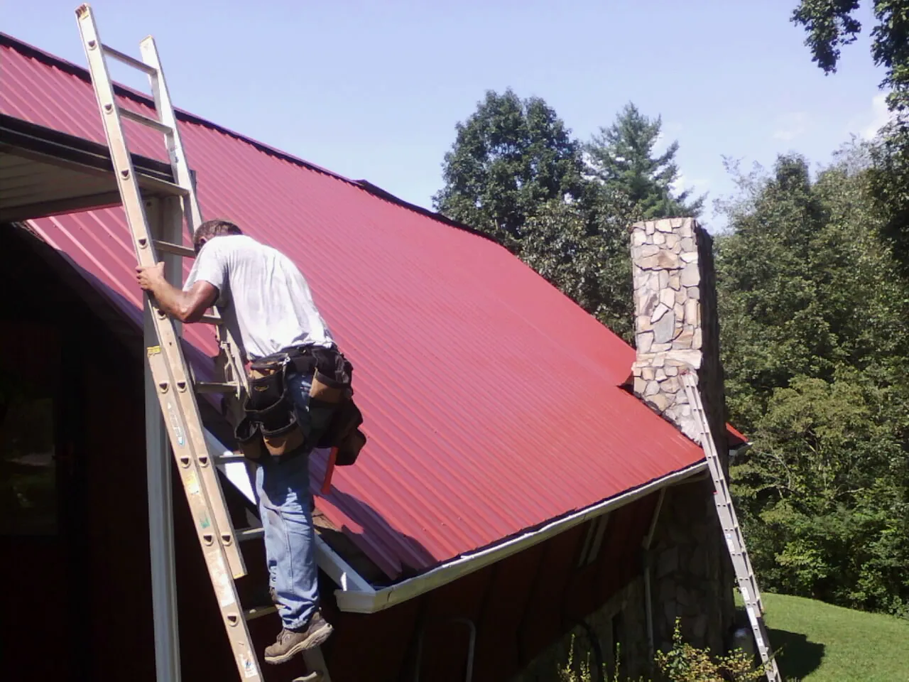Roofer on a ladder beside a red metal roof and stone chimney — Soffit & Fascia Repair by Roofing Brothers in Norwalk